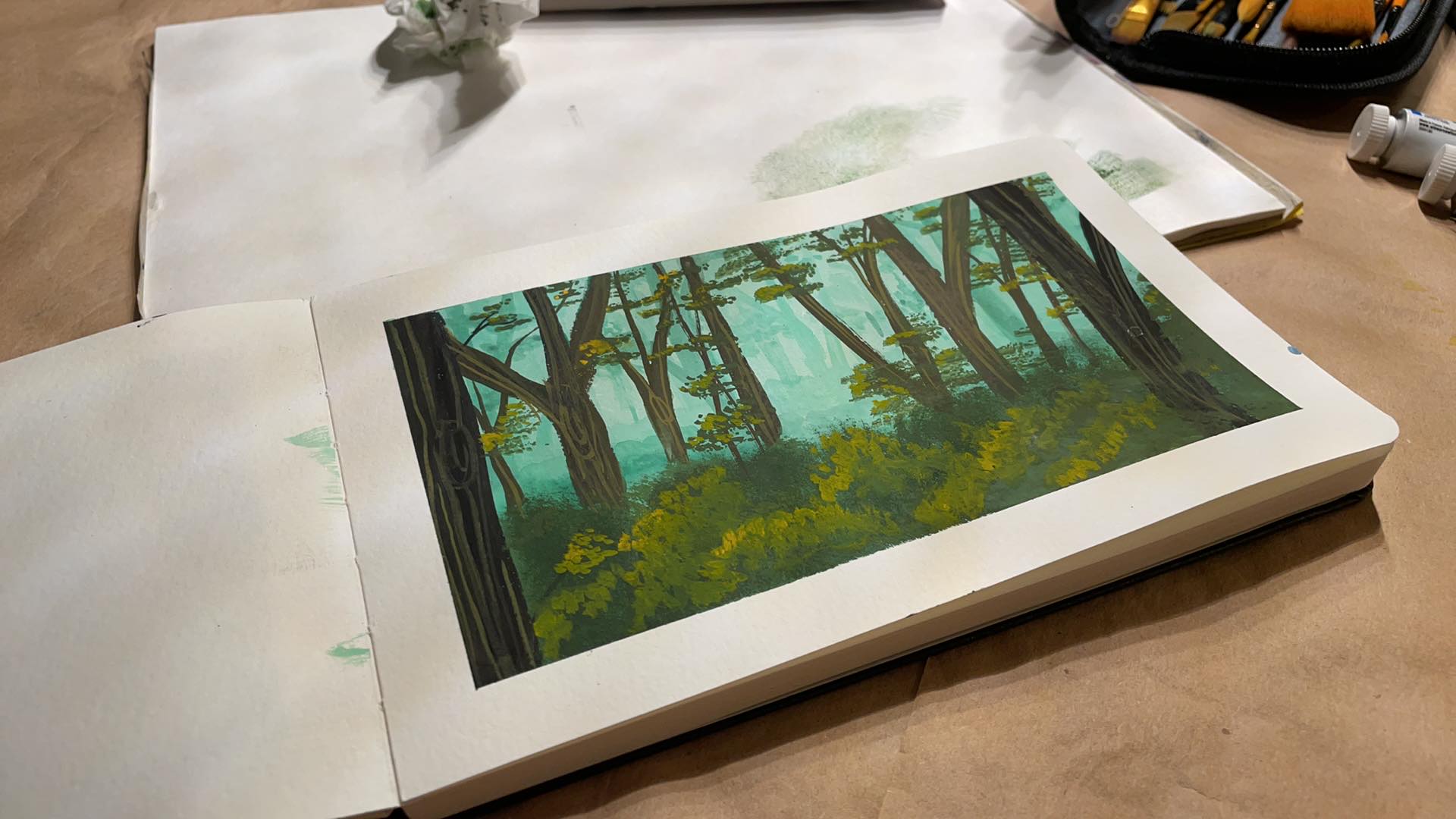 A gouache painting of a misty forest in teal and green, dense tree trunks silhouetted against glowing fog, painted in a sketchbook resting on a larger sheet with paint tubes and brushes nearby