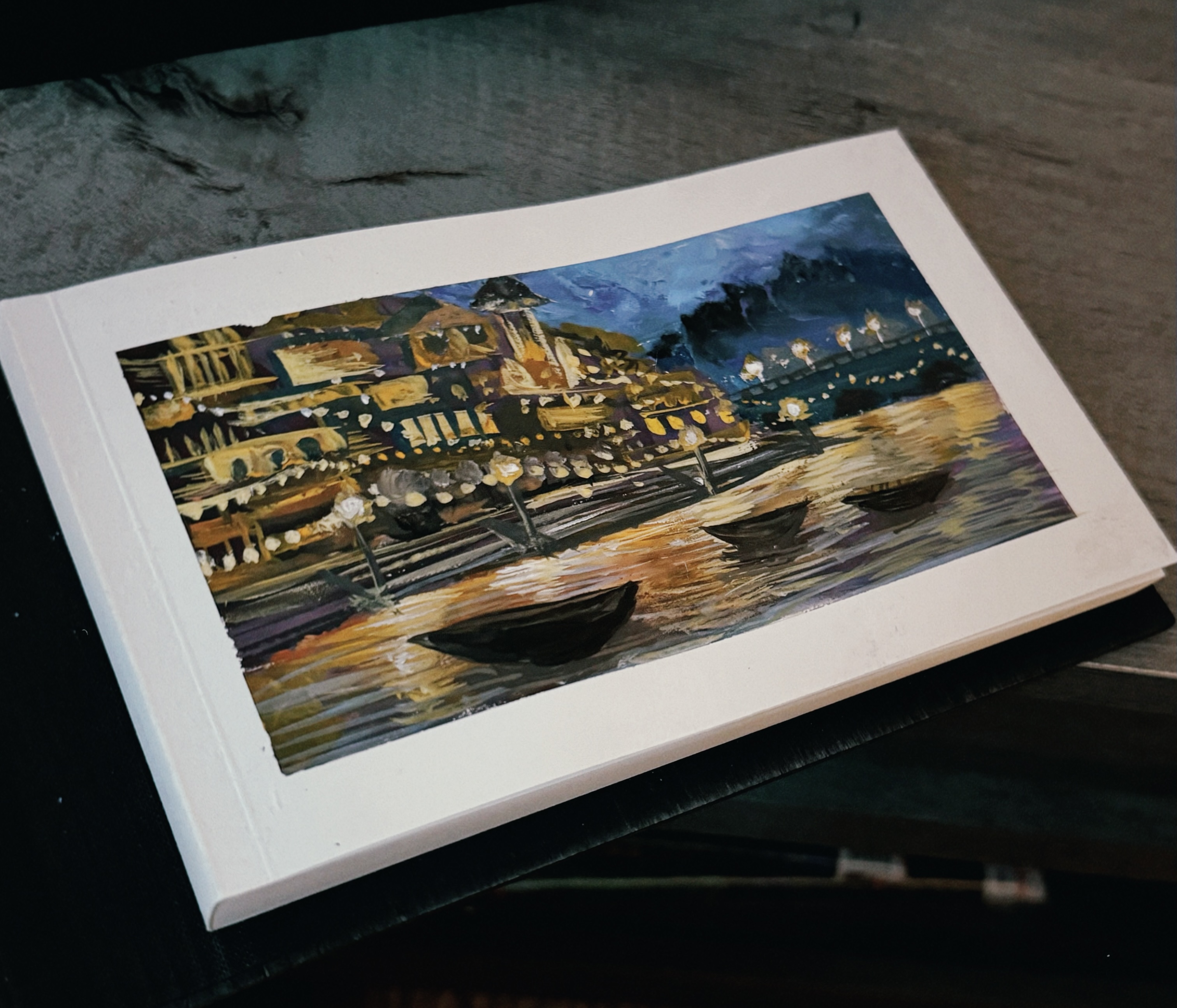 A painting of a riverside town glowing at night, string lights reflecting off the water beside small wooden boats moored along the bank
