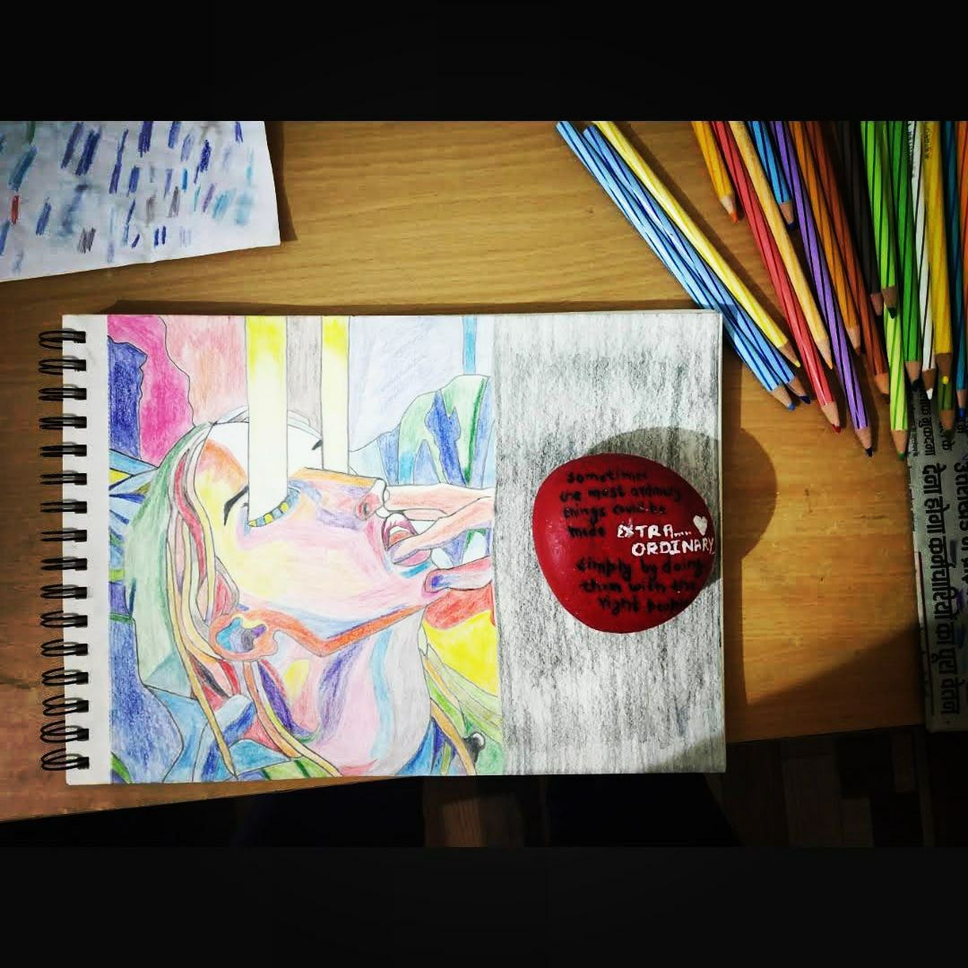A colour-pencil sketchbook spread of an abstract portrait with a face tilted back, mid-shout, rendered in overlapping washes of pink, blue, green, and orange, beside a painted red stone reading 'Sometimes the most ordinary things become extraordinary simply by doing them with the right heart'