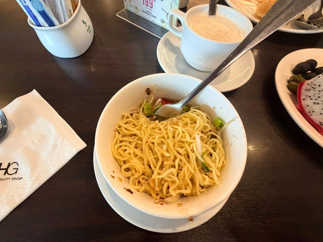 A bowl of spicy noodles next to a coffee at a hotel breakfast table