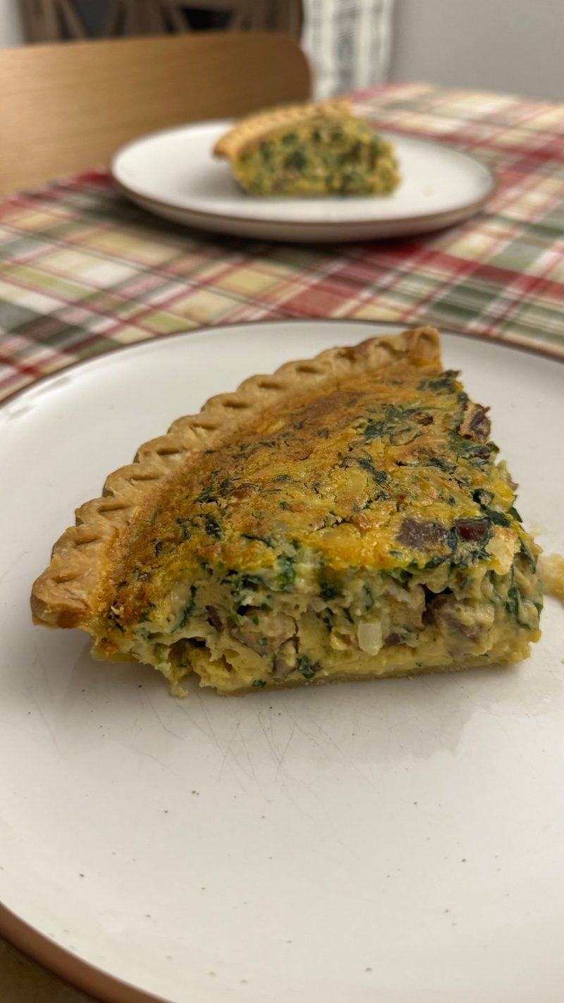 A slice of spinach and mushroom quiche on a plate, with a second slice in the background