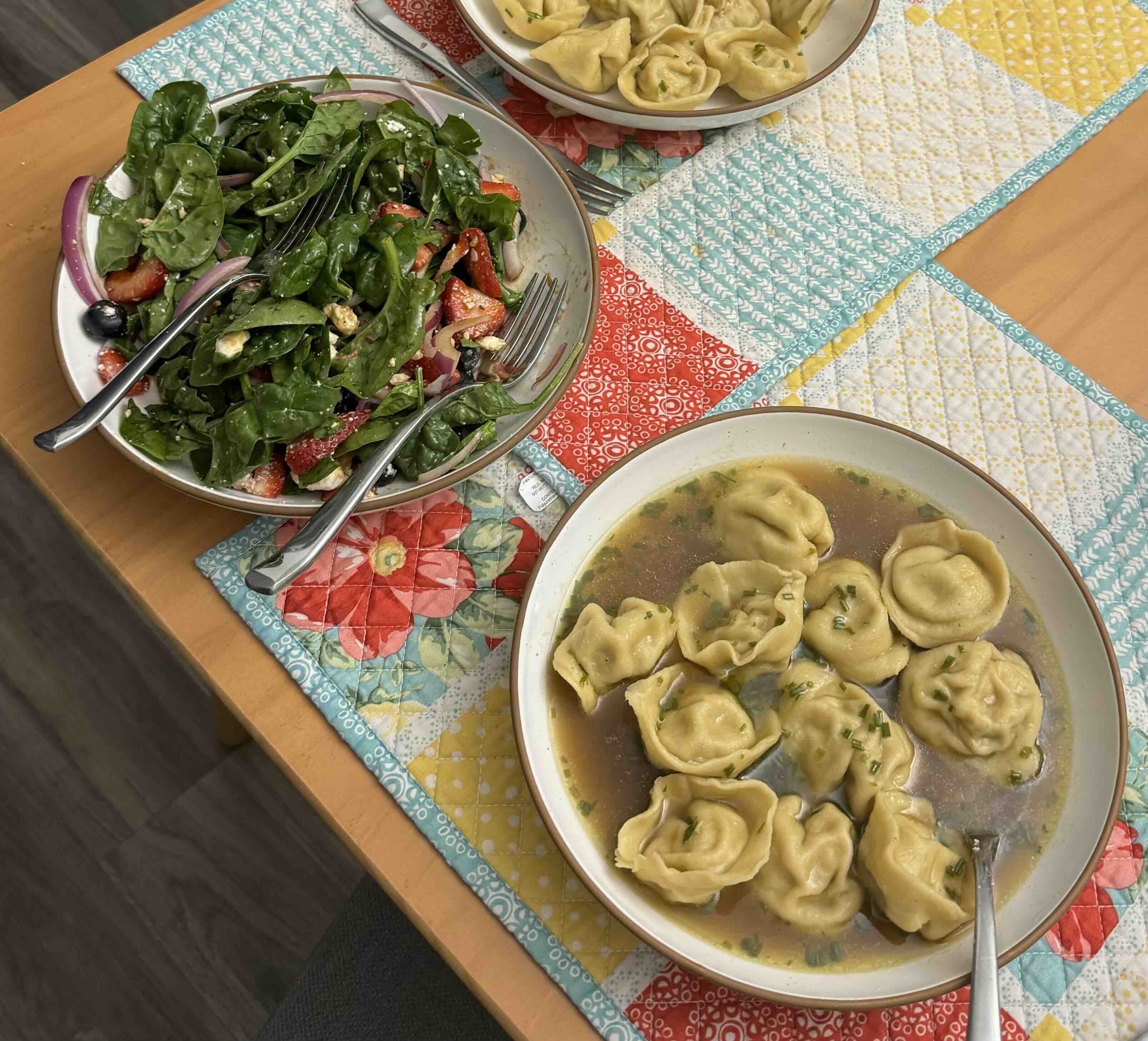 An overhead shot of a homemade meal on a quilted table runner: a spinach salad with strawberries, blueberries, red onion, and feta, alongside a bowl of dumplings in broth and a plate of dumplings on the side
