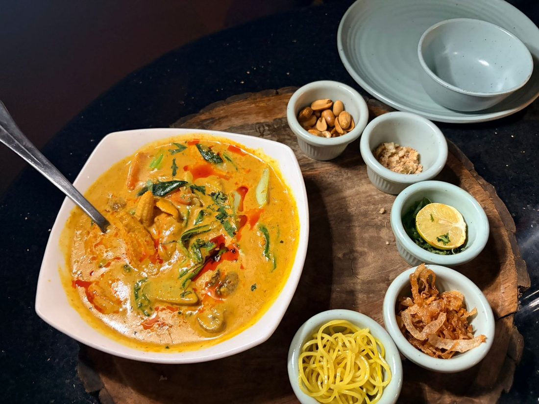 A bowl of yellow curry with baby corn and herbs, served with peanuts, crispy shallots, lime, and egg noodles on the side