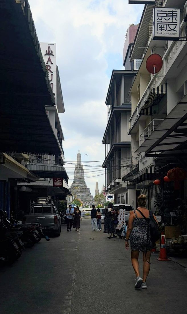 A narrow Bangkok street lined with shopfronts, with the spire of Wat Arun visible in the distance