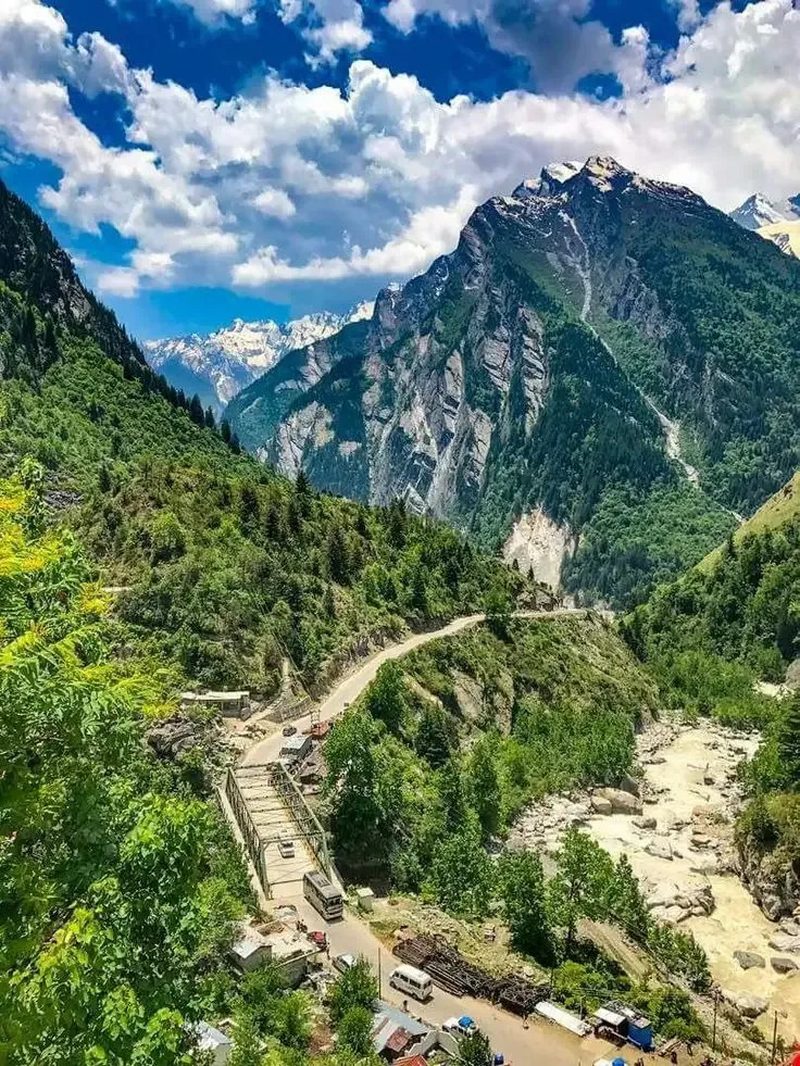 A winding mountain road through a green Himalayan valley, with a river and snow-capped peaks in the distance
