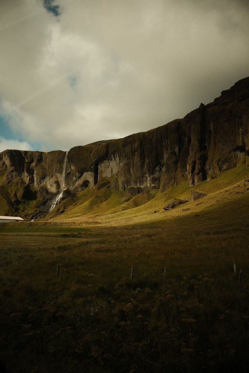 A waterfall dropping down a mossy basalt cliff over an Icelandic farm field