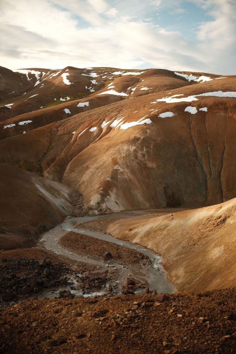 Rust-coloured volcanic highlands in Iceland streaked with snow, with a meltwater stream cutting through the valley